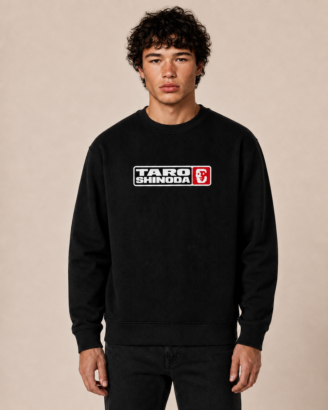 A man wearing a black crewneck sweatshirt with a white, black, and red 'TARO SHINODA' graphic on the chest, standing against a solid beige background.
