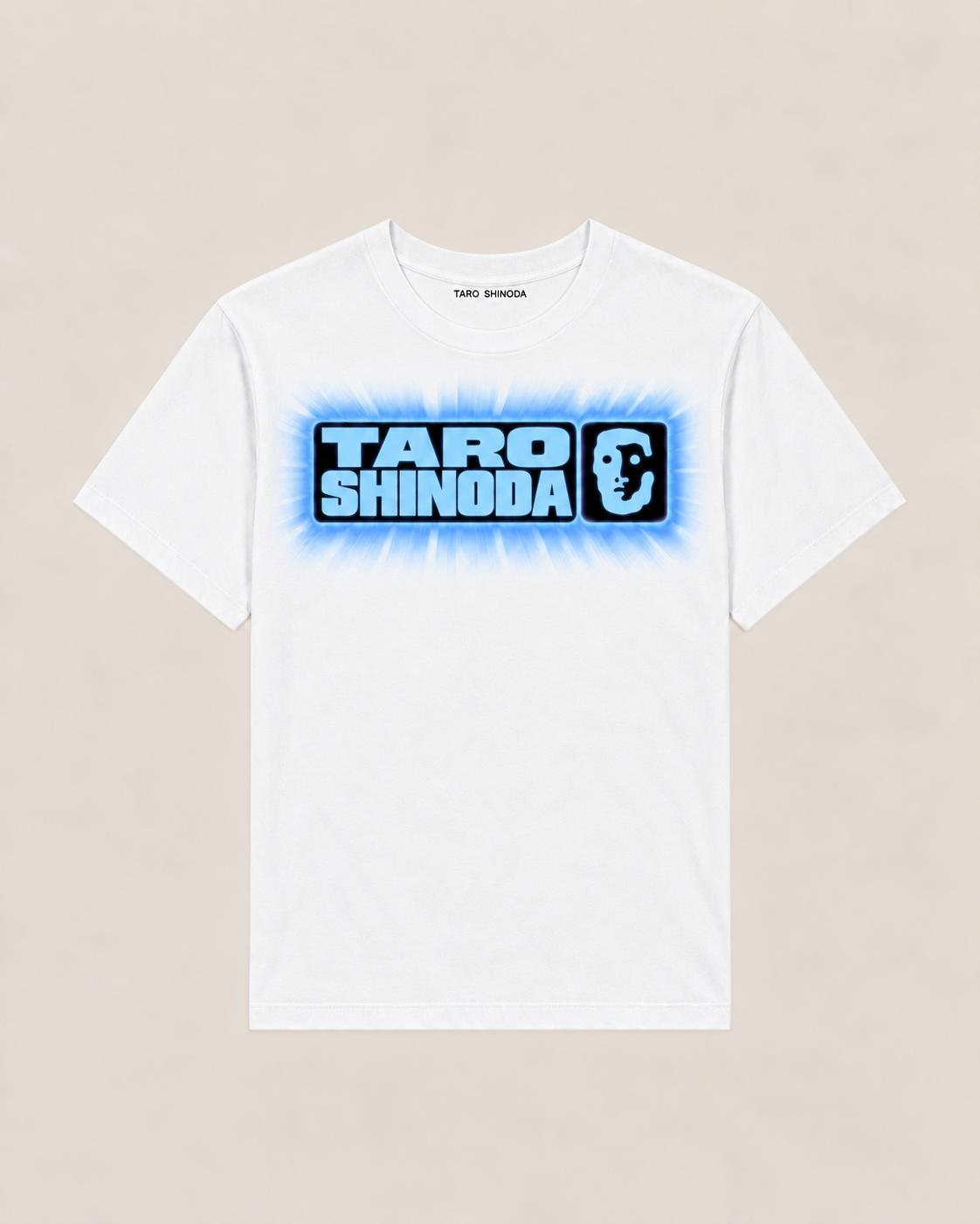 A white short-sleeve crewneck T-shirt featuring a blue and black graphic print on the chest that reads 'TARO SHINODA' with a stylized face logo, displayed flat against a light beige background.