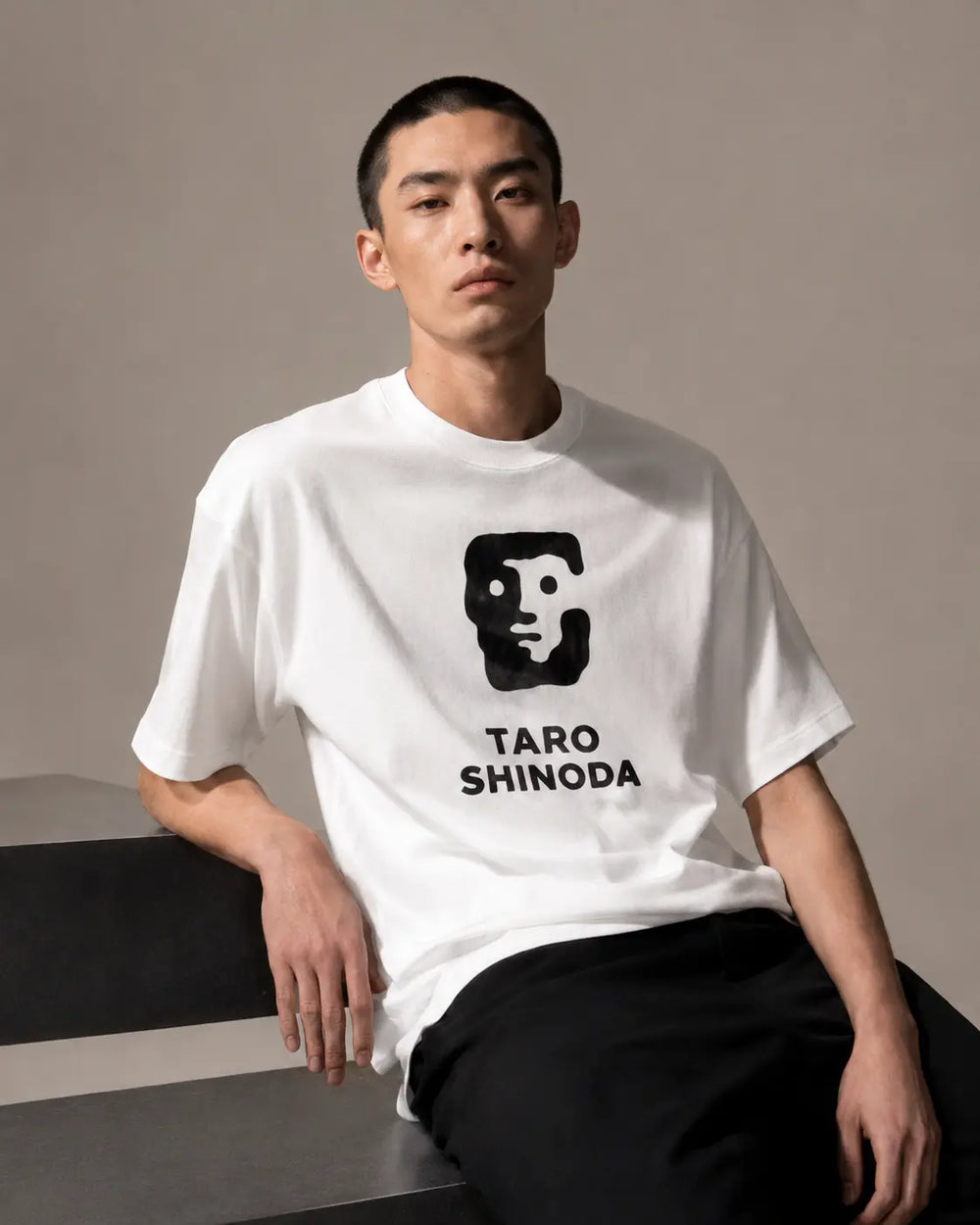 A man wearing a white T-shirt with a black graphic of a face and the text 'TARO SHINODA' printed on the front, paired with black pants, seated against a neutral beige background.