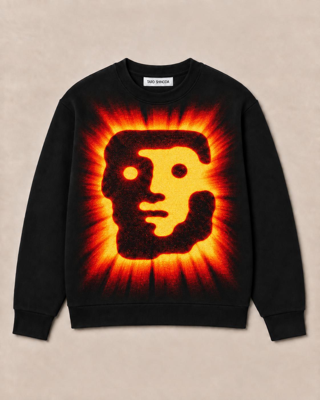 A black crewneck sweatshirt featuring a large, vibrant graphic of a stylized face in yellow and orange, surrounded by a fiery red and orange sunburst pattern, laid flat on a light beige background.