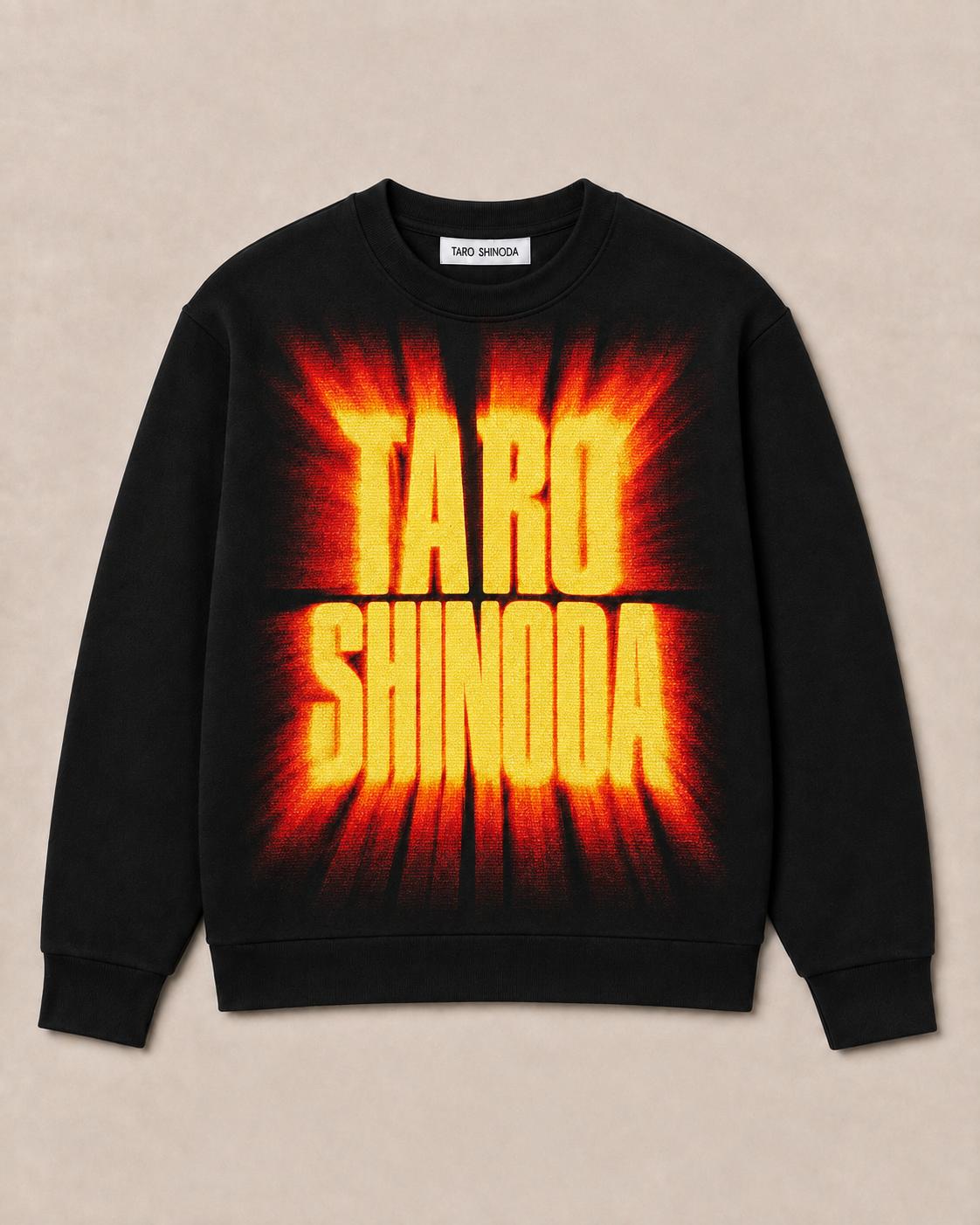 A black crewneck sweatshirt featuring a large graphic print of the name 'TARO SHINODA' in bold yellow letters with a red and orange flame-like burst background, displayed flat against a light beige textured backdrop.