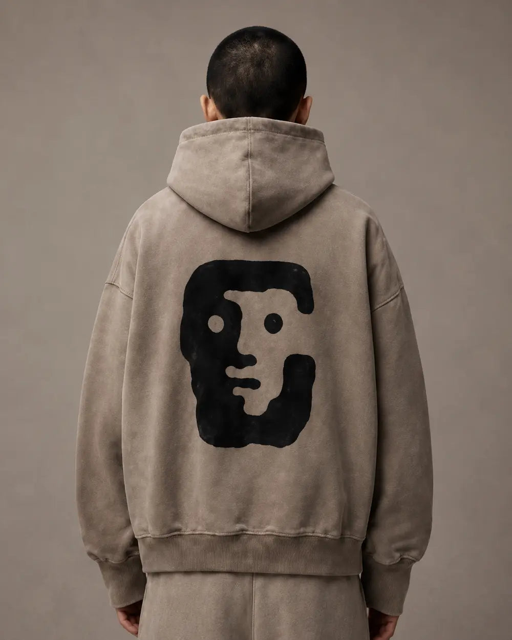 A person viewed from behind wearing a beige oversized hoodie with a large black face graphic printed on the back, against a plain taupe studio background.